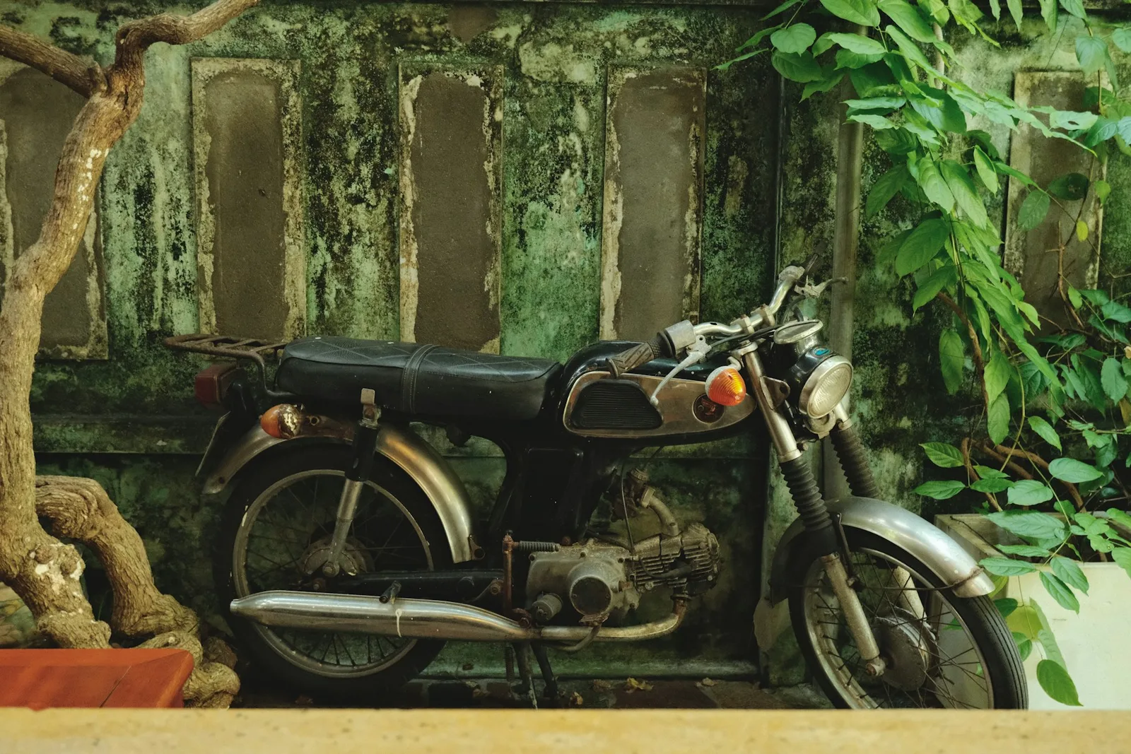 An old motorcycle with worn paint beside a moss-covered wall