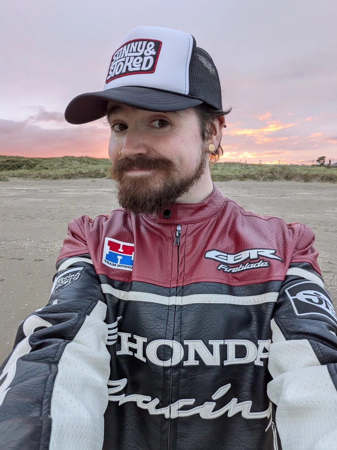 Jb wearing his burgundy, black and ivory Honda racing jacket and a trucker cap, with a sunset behind him
